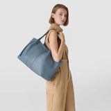 Side view: SECRET TOTE BAG IN CACHEMIRE LEATHER AND MOSAICO worn over the shoulder, indigo cachemire leather with Mosaico trim, model in a beige jumpsuit against a neutral background.