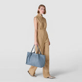 Front view: SECRET TOTE BAG IN CACHEMIRE LEATHER AND MOSAICO is carried at the side in the hand of a standing woman in a beige jumpsuit; light blue with a central Mosaico detail.