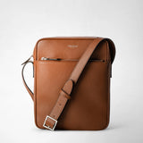 Frontal view: NORTH SOUTH CROSSBODY BAG IN CACHEMIRE LEATHER shows a flat brown crossbody bag in pebbled calfskin leather, adjustable shoulder strap worn diagonally, front zip pocket, 'SERAPIAN MILANO' lettering, neutral light gray background.