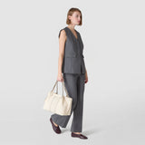 Side view: woman carrying SMALL SECRET TOTE BAG IN CACHEMIRE LEATHER AND MOSAICO in cream by its two short handles, standing in a gray outfit against a light, neutral background.