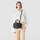 Frontal view: SMALL MARO BAG IN CACHEMIRE LEATHER AND MOSAICO, black handbag worn crossbody by a model against a neutral background, two handles, nappa leather strips knotted at the sides. Inscription: 'SERAPIAN'.