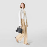 Front view: SMALL MARO BAG IN CACHEMIRE LEATHER AND MOSAICO is carried in the hand of a standing woman, black Mosaico details and knotted nappa leather strips, light neutral background.