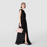 Side view: A woman in a black sleeveless evening dress carries the SMALL MARO BAG IN CACHEMIRE LEATHER AND MOSAICO in Antique Rose by its short handles against a neutral light gray background.