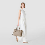 Frontal view: MEDIUM MARO BAG IN CACHEMIRE LEATHER AND MOSAICO – model carries Sahara-colored bag by the handles, relaxed at their side, against a light studio background.