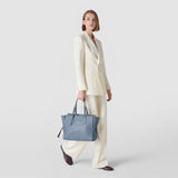 Front view: MEDIUM MARO BAG IN CACHEMIRE LEATHER AND MOSAICO in indigo-blue Mosaico Cachemire leather, held by a woman in a cream pantsuit by its two handles.