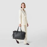 Side view: A woman carries the MAXI MARO BAG IN CACHEMIRE LEATHER AND MOSAICO on her arm; large black tote with a Mosaico panel at the top, supple Cachemire leather and side knots against a light background.