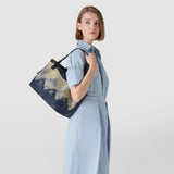 Frontal view: Woman wears SMALL SECRET TOTE BAG IN DENIM AND RAFFIA over her shoulder; navy and light-gold jacquard raffia pattern, narrow handles, visible 'SERAPIAN' script.