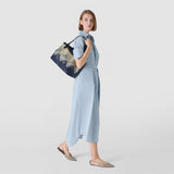 Side view. SMALL SECRET TOTE BAG IN DENIM AND RAFFIA worn over the shoulder; navy–light-beige jacquard raffia pattern, woman in a light-blue midi dress against a neutral background.