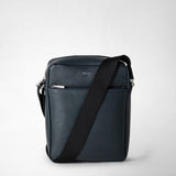 Front view: NORTH SOUTH CROSSBODY BAG IN EVOLUZIONE LEATHER shows a flat navy blue crossbody bag in textured calfskin leather with a front zip pocket and a wide black adjustable shoulder strap against a light background. Text: 'SERAPIAN MILANO'.