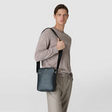Frontal view: Man wears the NORTH SOUTH CROSSBODY BAG IN EVOLUZIONE LEATHER over his shoulder; compact, rectangular dark green leather bag with visible zipper and 'SERAPIAN MILANO' print, neutral light background.