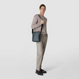 Front view: NORTH SOUTH CROSSBODY BAG IN EVOLUZIONE LEATHER hangs crossbody over the shoulder of a standing man, compact, smooth dark leather, subtle zipper, neutral light gray background.