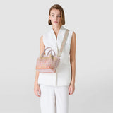 Frontal view: Woman wears MINI SECRET BAG IN MOSAICO MESTIERI D'ARTE over a white outfit; small trapezoid handbag in Bokashi pink with mosaic pattern, short handles and wide shoulder strap.
