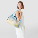 Frontal view: woman wearing SECRET TOTE BAG IN MOSAICO MESTIERI D'ARTE over her shoulder; pastel blue-and-yellow geometric woven pattern, white outfit, neutral studio background.
