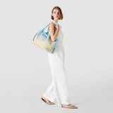 Side view: A woman wears the SECRET TOTE BAG IN MOSAICO MESTIERI D'ARTE casually over her shoulder; large bokashi azure-blue and yellow tote with woven pattern, white outfit, neutral background.