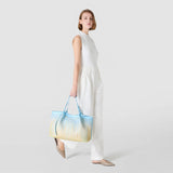 Side view — Model wears SECRET TOTE BAG IN MOSAICO MESTIERI D'ARTE in Bokashi Azure, holding it by the handles alongside a white, elegant outfit; woven gradient pattern.