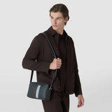 Front view: A man wears the CAMERA CROSSBODY BAG IN STEPAN over his shoulder. The dark patterned bag features a light blue central stripe and leather details.